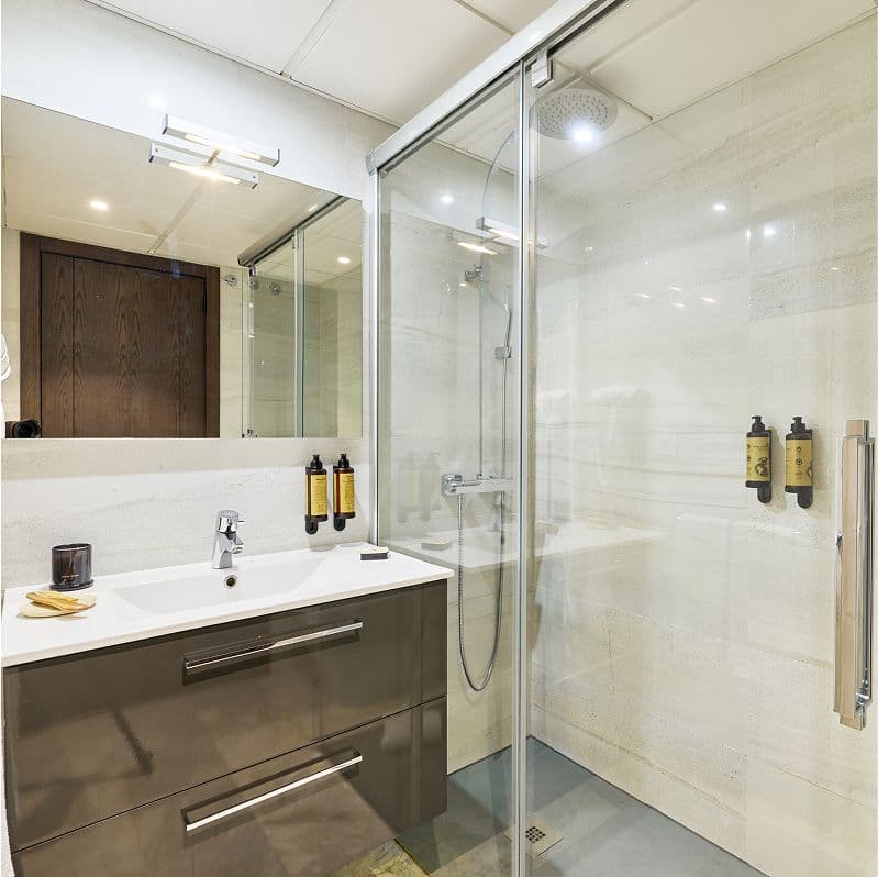 Baño moderno con mampara de vidrio, ducha efecto lluvia empotrada y mueble de lavabo gris con espejos y dispensadores de jabón de pared