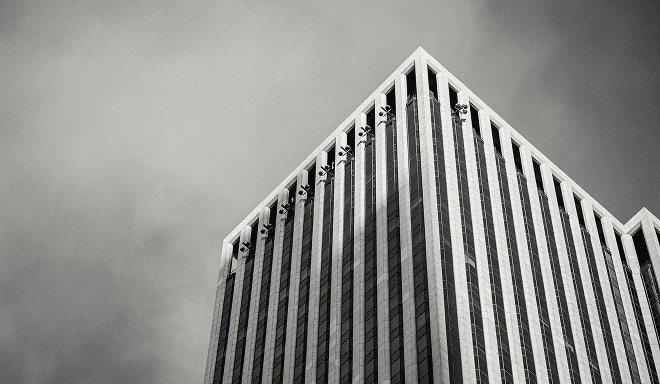 Fotografía en blanco y negro de un edificio de oficinas moderno de gran altura visto desde la calle, con líneas verticales y cielo nublado
