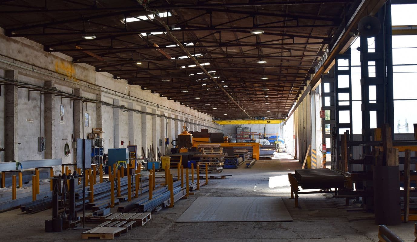 Pasillo central de nave industrial con vigas metálicas en el techo, materiales apilados y luz natural