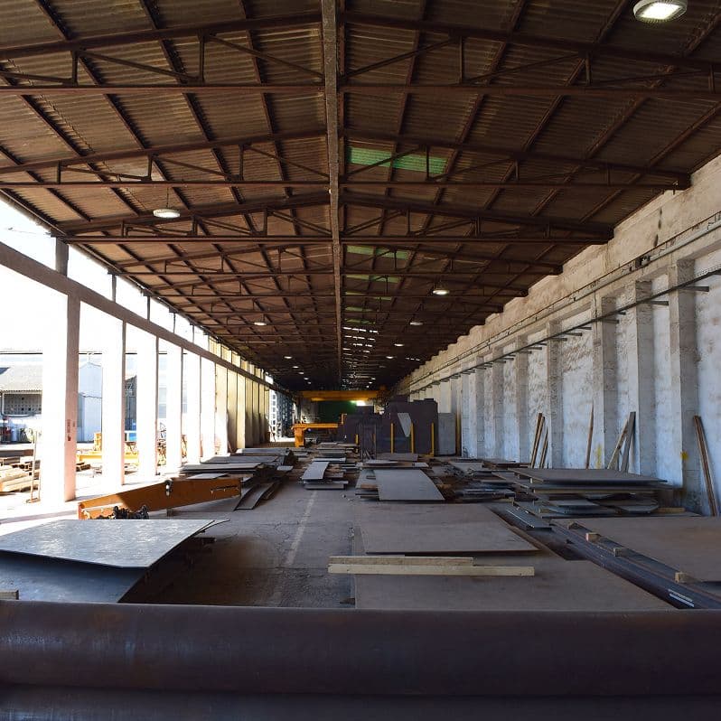 Detalle lateral de nave industrial con estructura metálica, suelo de hormigón y acumulación de perfiles de acero