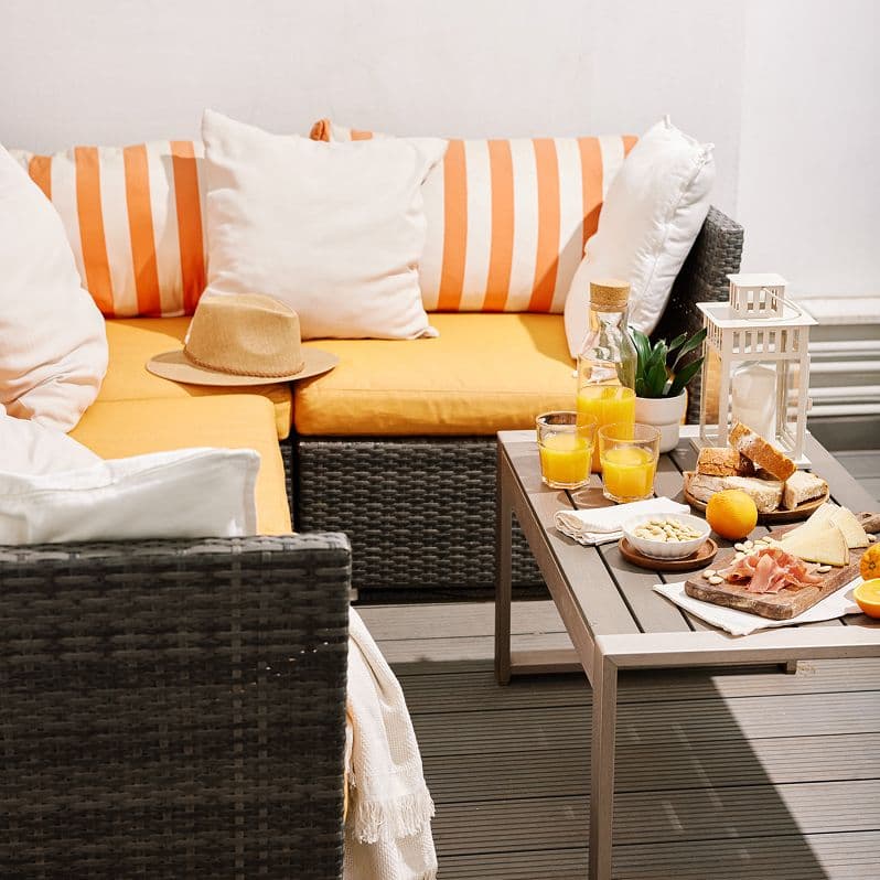 Zona de terraza con sofá de mimbre y cojines naranjas, mesa auxiliar con jugo de naranja, tabla de quesos y embutidos